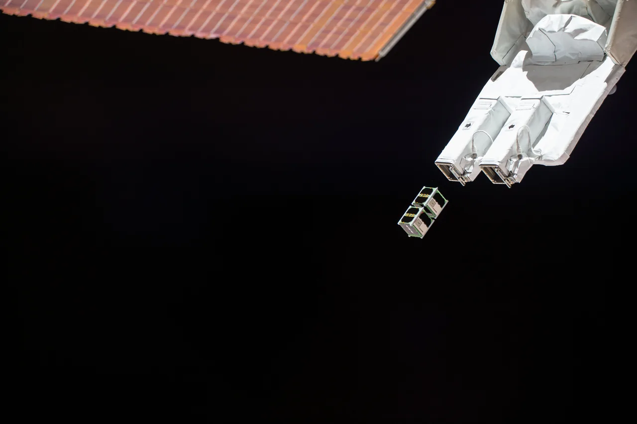 Two small CubeSats move away from a white deployer beneath a station solar array against dark space