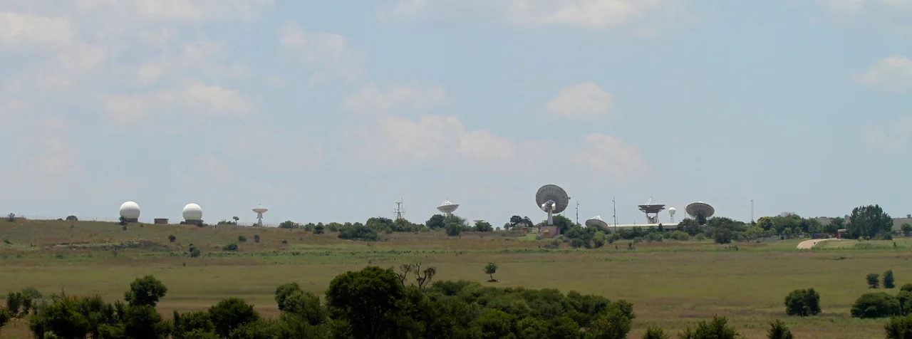 Several dish antennas and white radomes stand across a grassy ridge beneath a wide cloudy sky
