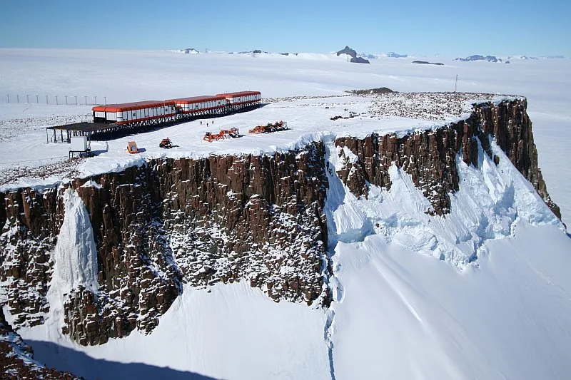 An orange-roofed research station stands on exposed dark rock surrounded by an extensive Antarctic ice landscape