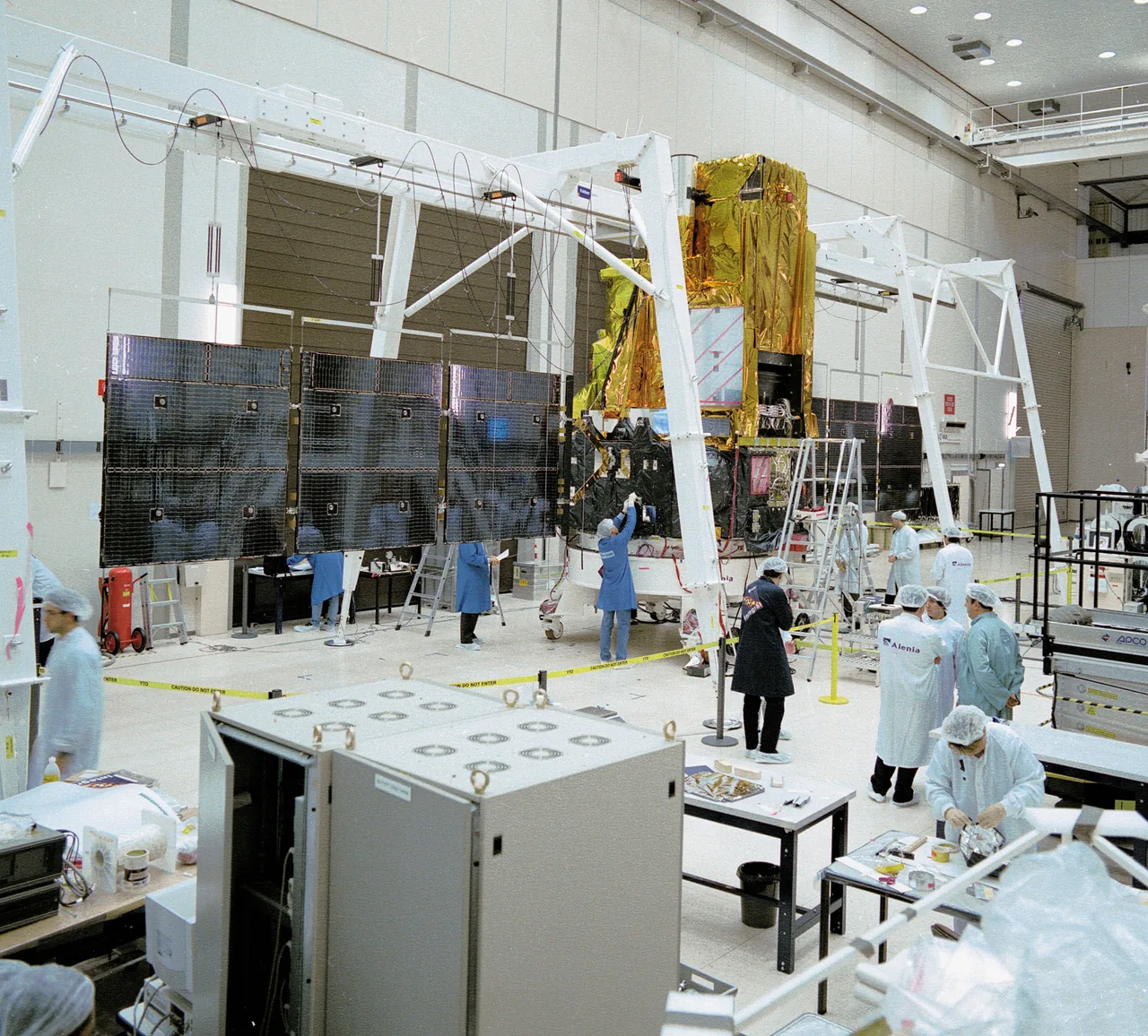 INTEGRAL’s tall gold-covered spacecraft body stands between extended dark solar arrays inside a cleanroom, with technicians nearby