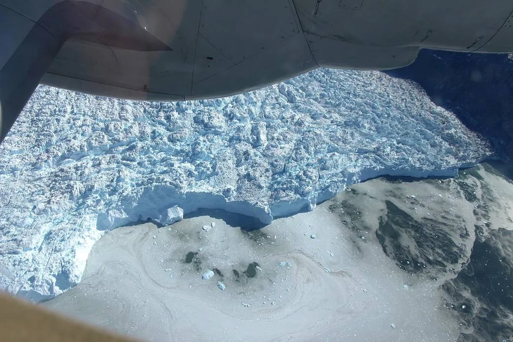 Rough crevassed glacier ice meets a front with separated ice pieces, viewed below an aircraft’s engine and landing-gear fairing.