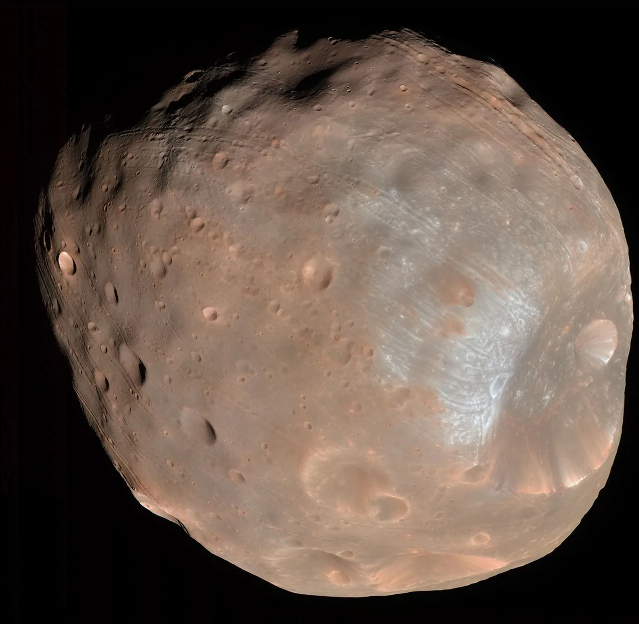 An irregular tan Phobos fills the frame, with Stickney crater at lower right, smaller craters, grooves, and bluish patches near the large crater.