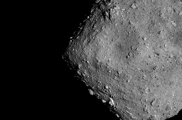 Close grayscale view of part of Ryugu, showing rough terrain, many boulders, a curved sunlit edge, and deep shadow at left.