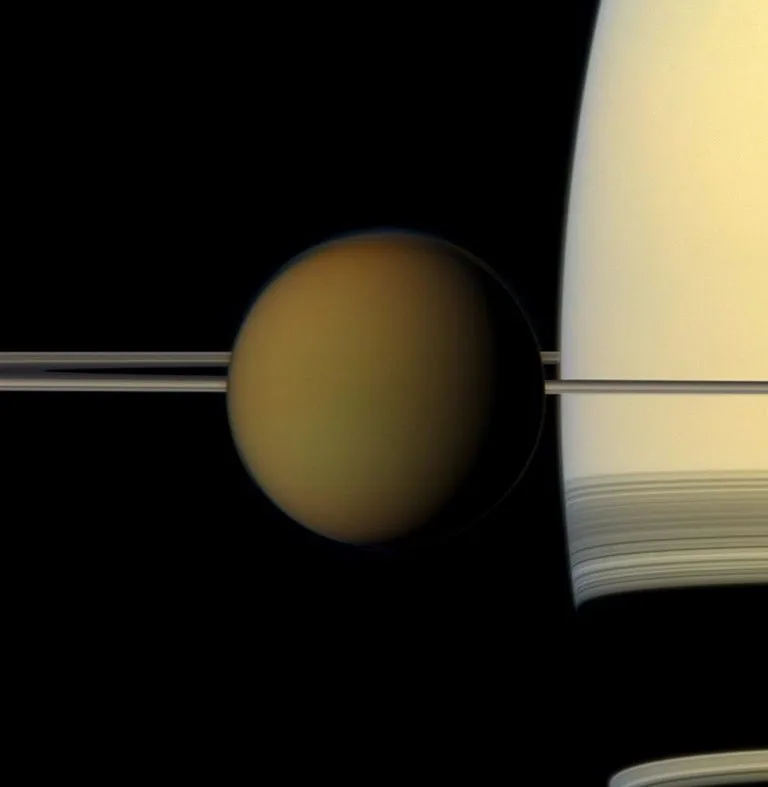 Hazy amber Titan appears in front of Saturn, whose pale disk and nearly edge-on rings extend across the background.