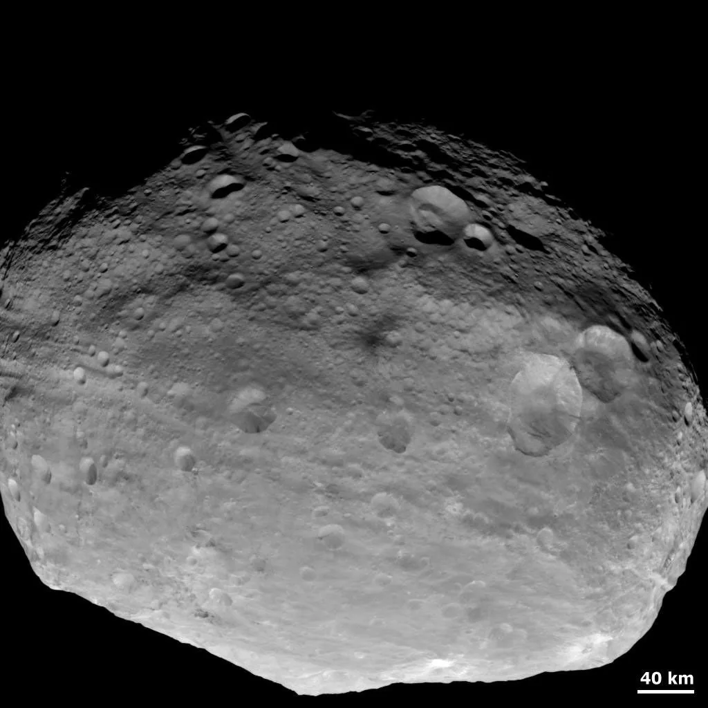 A grayscale Dawn view shows Vesta’s irregular outline, cratered surface, deep shadows near the top, and a 40-kilometer scale bar.