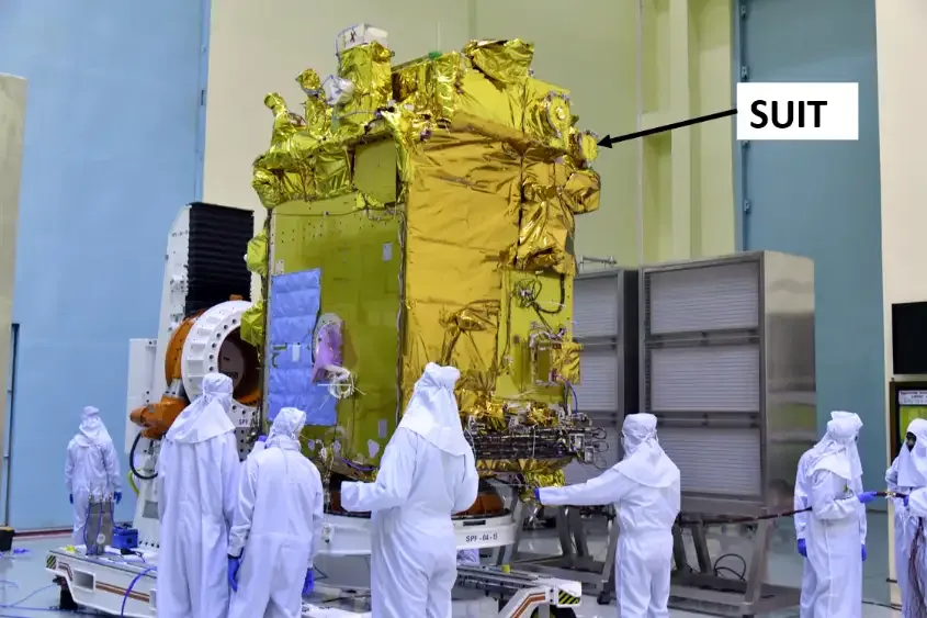 A tall gold-insulated Aditya-L1 spacecraft stands among cleanroom technicians; the source’s black arrow and SUIT label point to the telescope location.