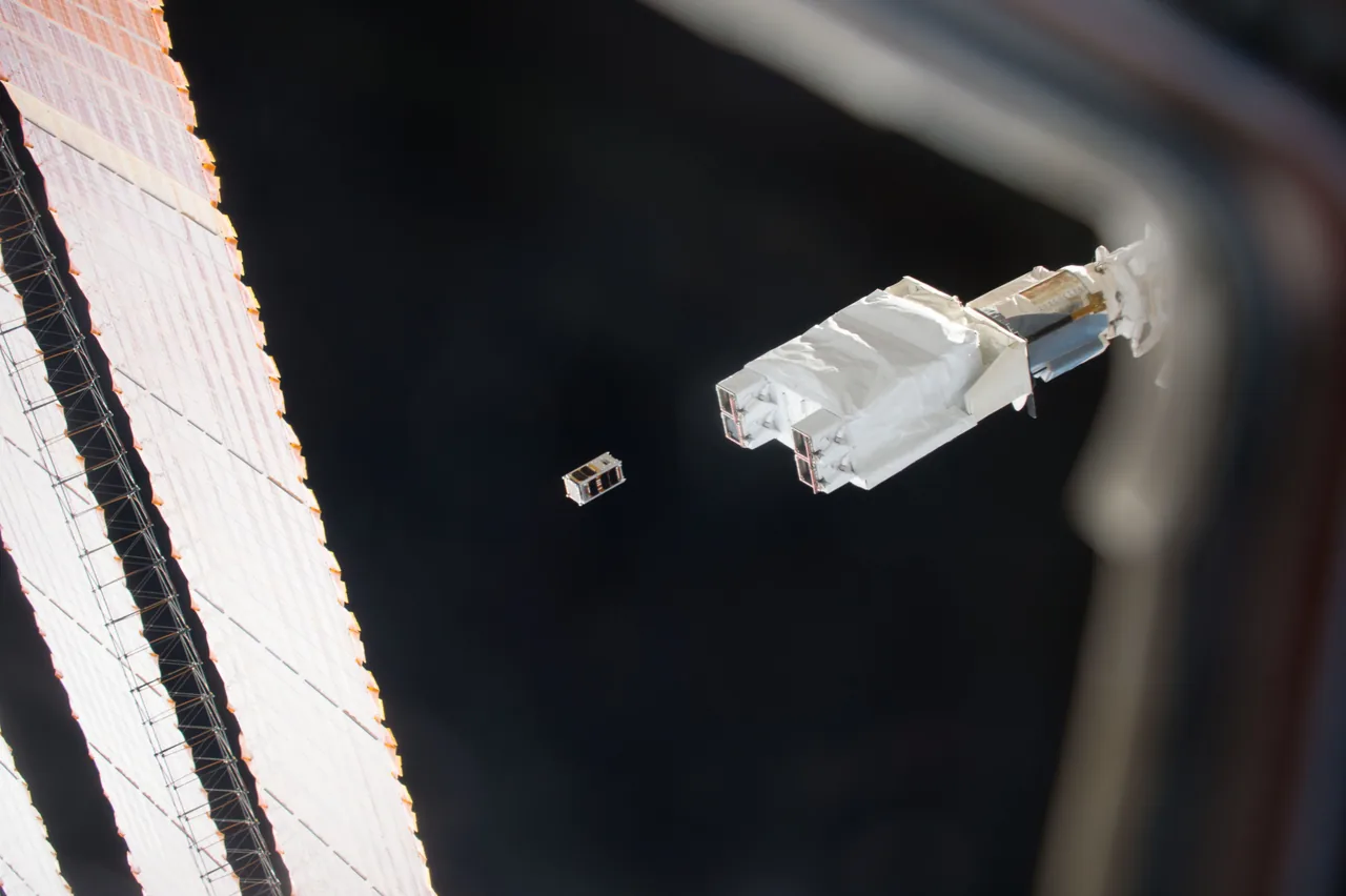 A small box-shaped satellite floats beside white deployment hardware and a station solar array against black space
