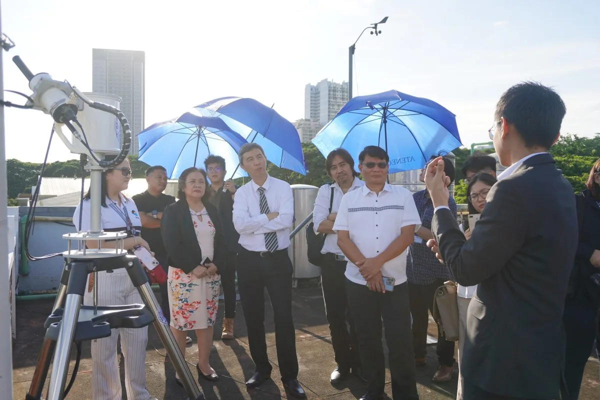 Visitors stand beside a tripod-mounted sky-viewing instrument on a sunny roof deck, with buildings behind them