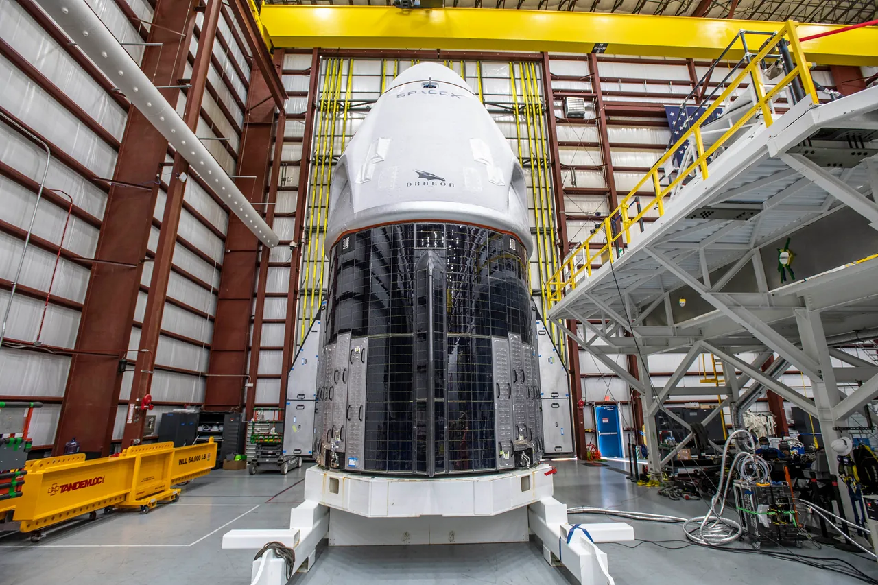Crew Dragon capsule and trunk standing vertically inside a launch processing building beneath yellow overhead cranes.