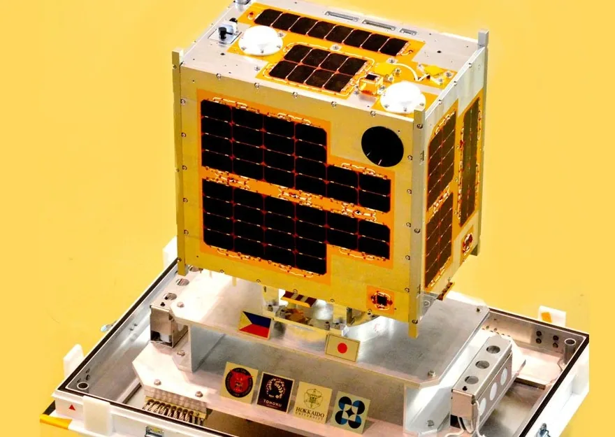 A compact box-shaped satellite with dark panels and circular instrument apertures sits on a mounting fixture