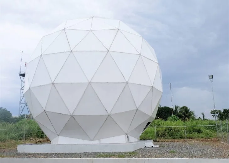 A white faceted radome stands on a concrete base beside a grassy field under an overcast sky
