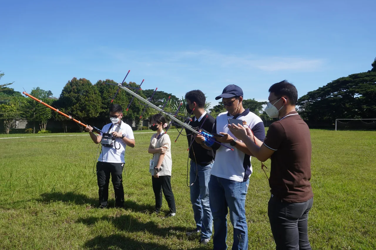 Five participants stand in a grassy field with directional antennas, radios and a phone