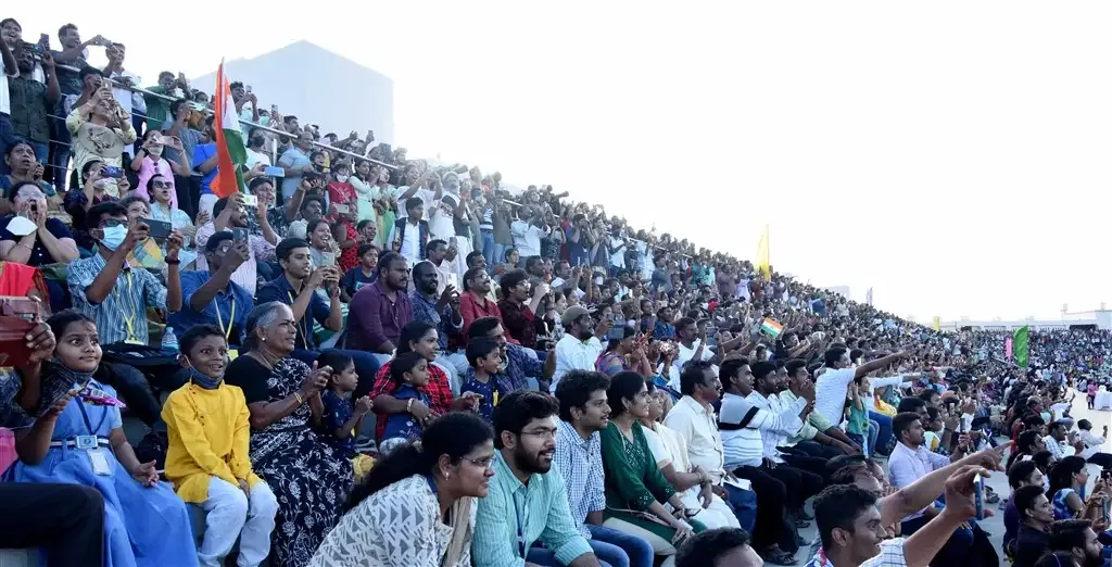 A crowded outdoor viewing gallery with adults and children watching and holding phones, with Indian flags among the audience
