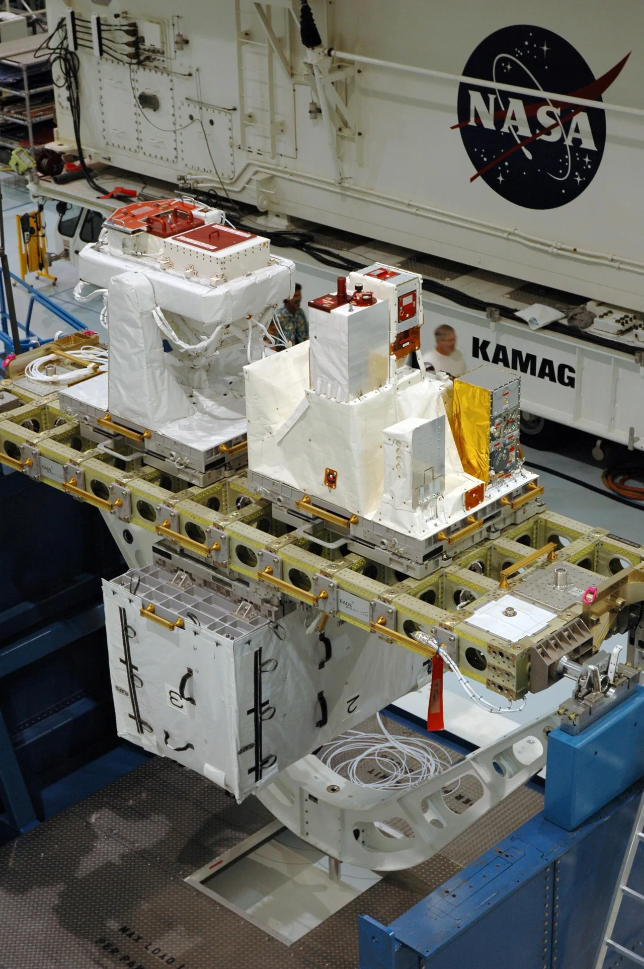 White-covered Columbus external payload hardware sits on a carrier surrounded by technicians and access platforms