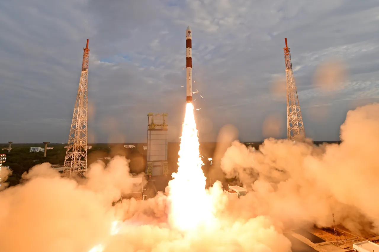 A white and red PSLV rocket rises between towers above a bright exhaust plume and clouds of smoke