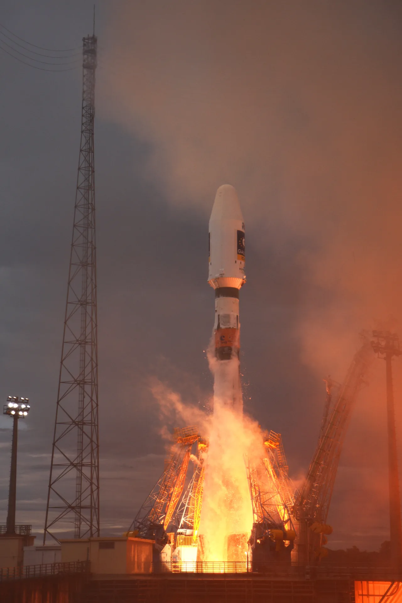 Soyuz rocket rising above the launch support arms in orange flame and smoke at the Guiana Space Centre.