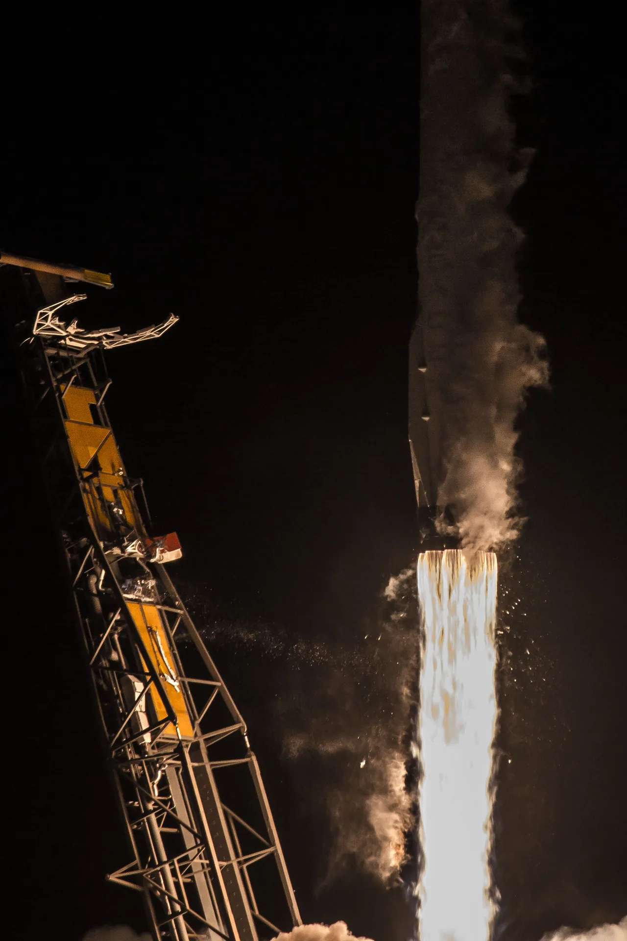 Night photograph of Falcon 9 rising beside a launch tower, with bright engine exhaust and vapor along the rocket