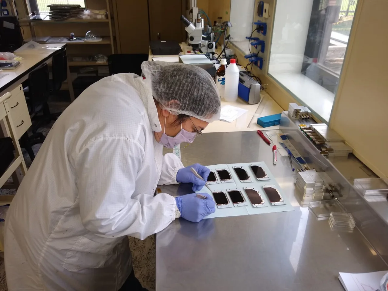 A masked technician wearing gloves and a hair covering works on small dark solar-cell panels at a laboratory bench