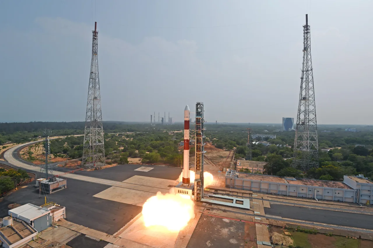 A white and red PSLV rocket lifts off beside its service structure between tall lightning towers in daylight