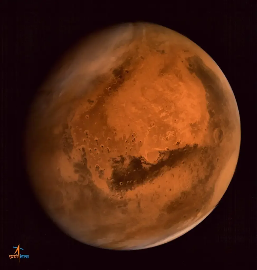The orange-red disc of Mars fills most of a dark frame, with bright limb haze, cratered terrain and a dark elongated surface region; the original ISRO mark remains at lower left.