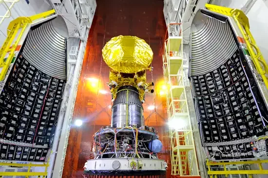 A gold-covered spacecraft sits above its launch stage between two open payload-fairing halves in a brightly lit integration tower.