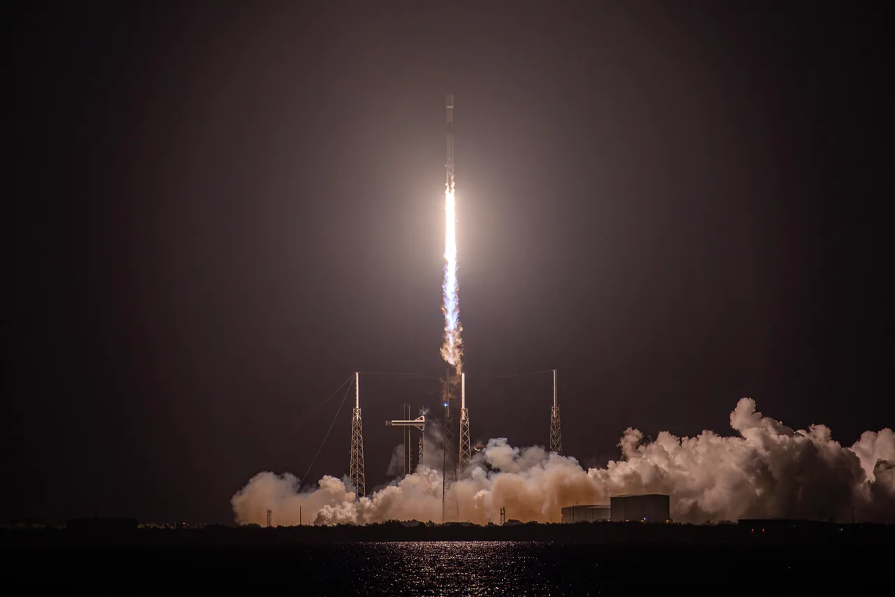 A Falcon 9 rises above a brightly illuminated exhaust plume and clouds at a nighttime launch pad