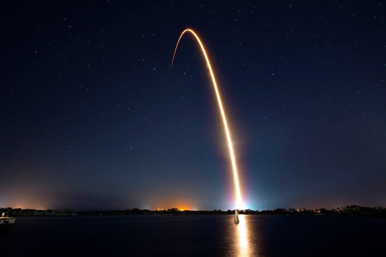 A long bright curved launch trail rises across a dark starry sky above water reflecting the light