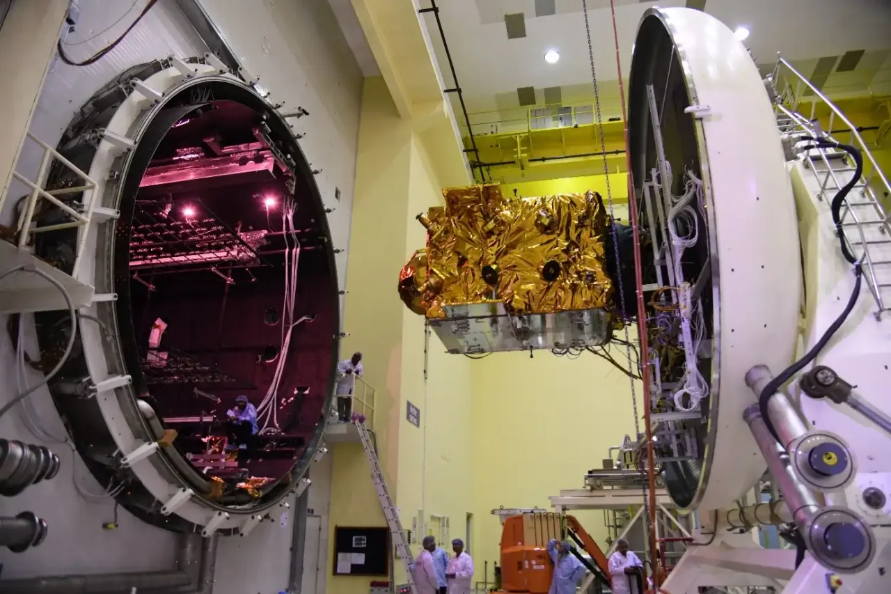 The gold-insulated NVS-02 spacecraft hangs beside the open circular doorway of a large thermal-vacuum chamber, with technicians and test equipment below.