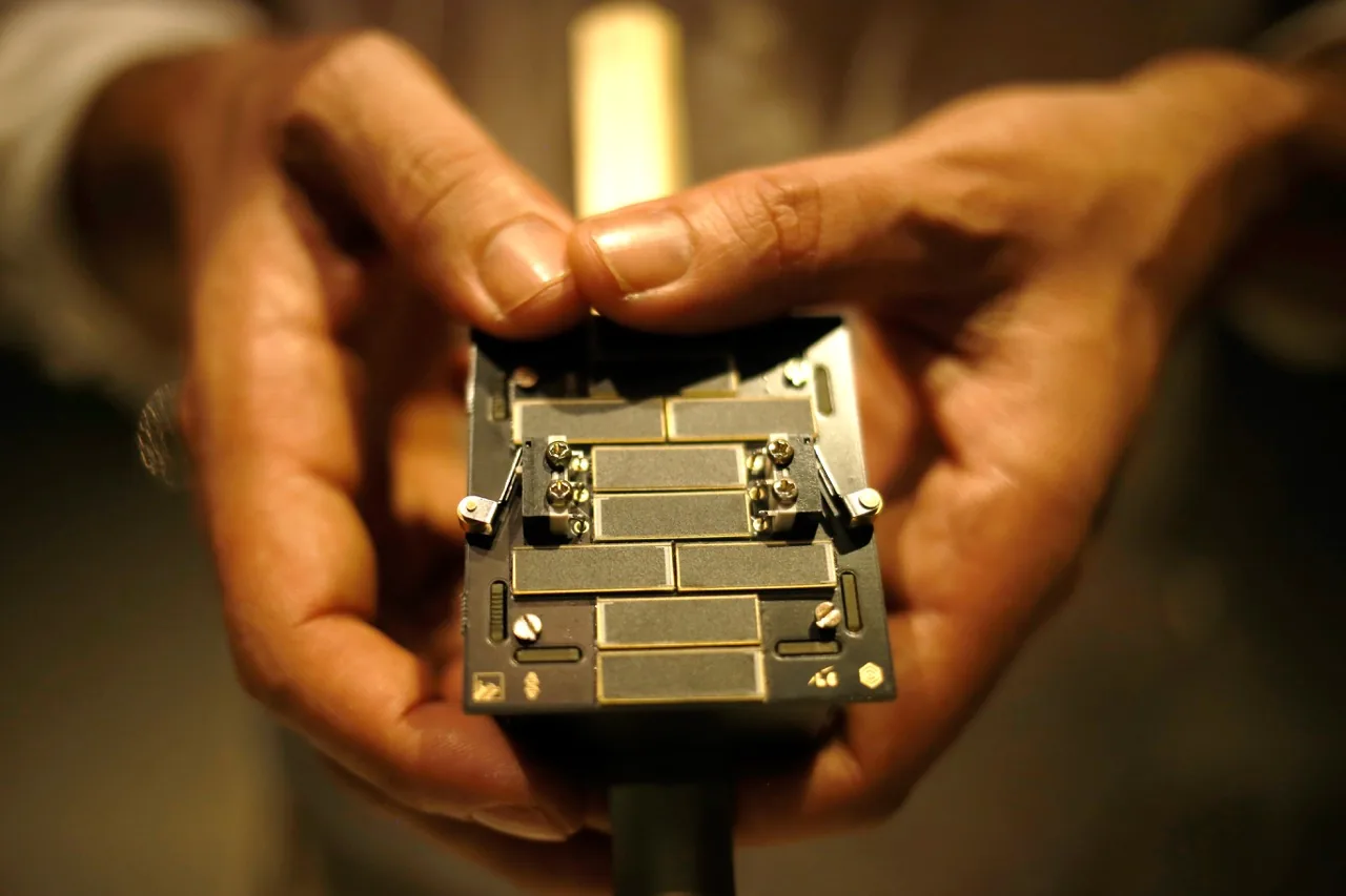 Hands hold the small Orbital Temple project satellite, showing its circuit boards and outer surfaces
