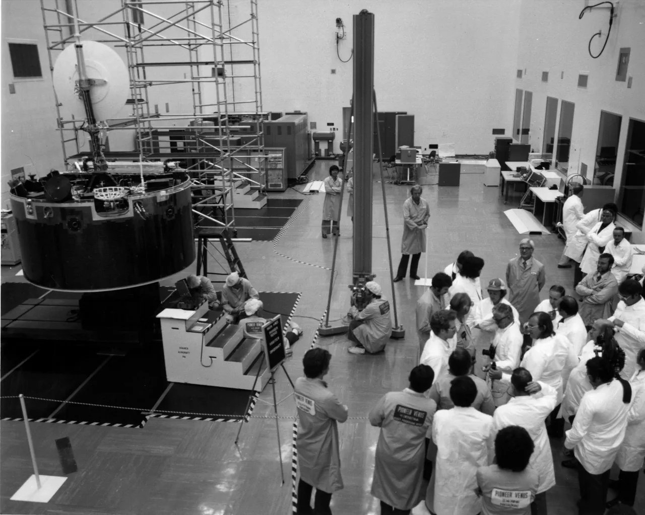 People gather around a cylindrical spacecraft with a dish antenna in a high-bay room