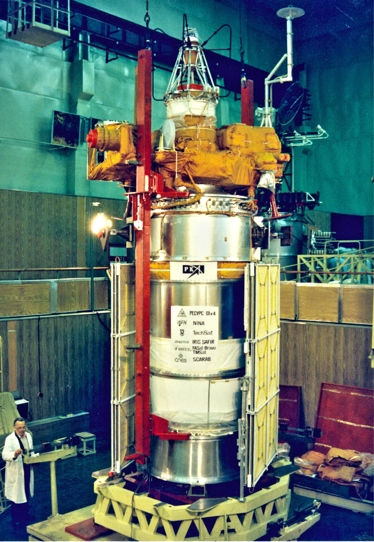 A tall silver and gold spacecraft stands vertically inside a preparation building, partly obscured by red supports; its base is labeled Resurs-O1 No. 4.