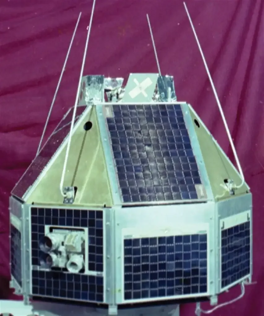 A faceted satellite with solar-cell panels, projecting antennas and optical apertures stands against a purple fabric backdrop.