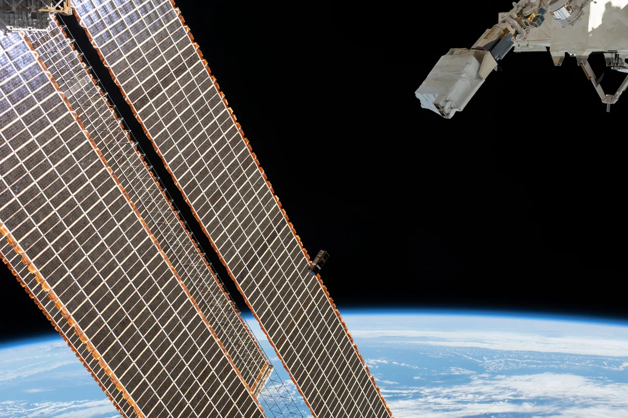 A small dark CubeSat passes beside large station solar arrays, with Earth below and deployment hardware above