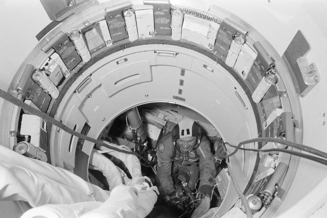 Astronauts stand beside the cylindrical Apollo–Soyuz docking module in a ground processing facility.