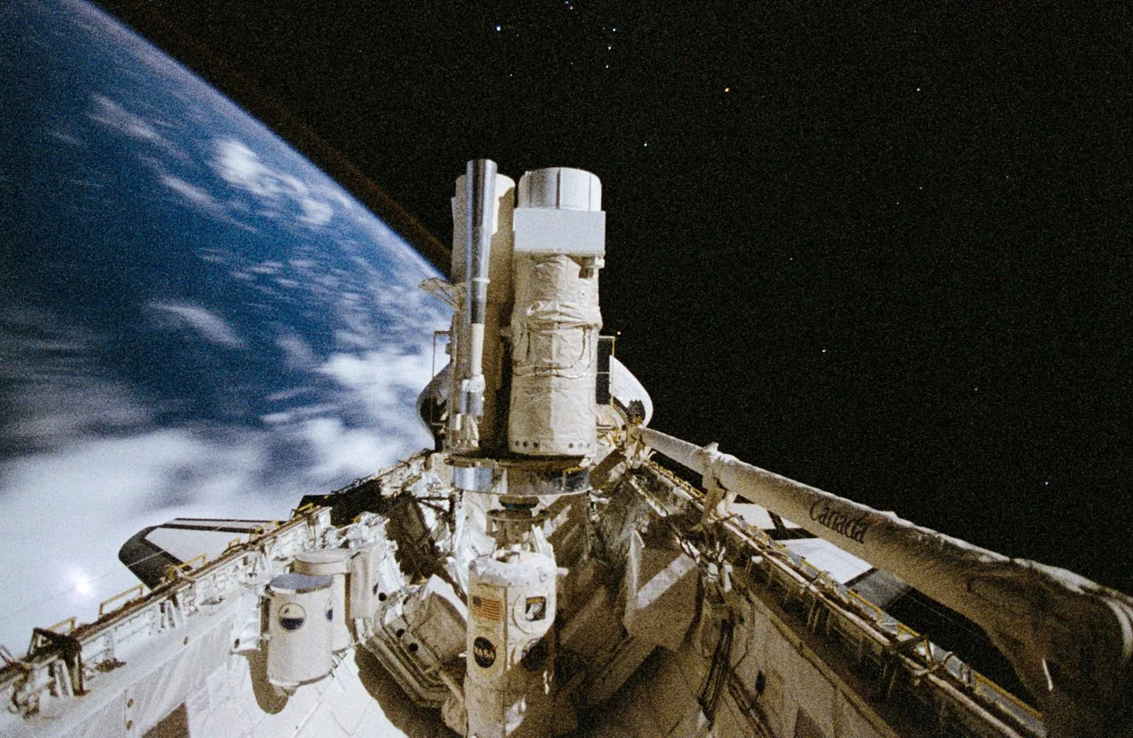 Wrapped telescope hardware fills Endeavour’s cargo bay, with a blue Earth edge and faint star points beyond.