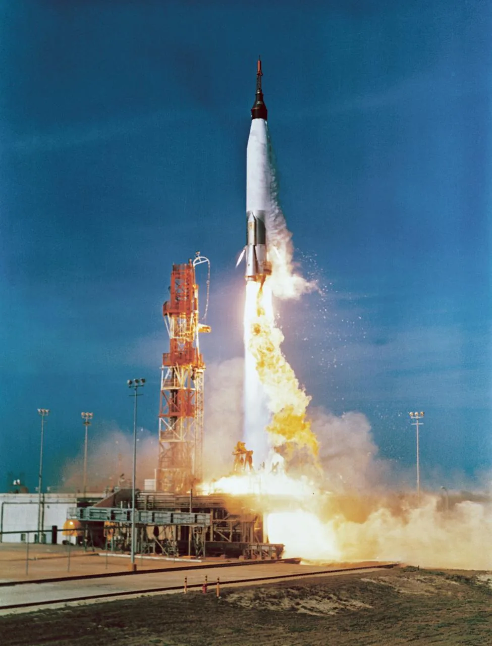 An early Atlas rocket rises above its launch platform with bright exhaust against a blue sky
