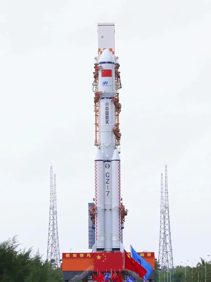 White Chang Zheng 7 rocket with four boosters upright on an orange mobile platform under an overcast sky.