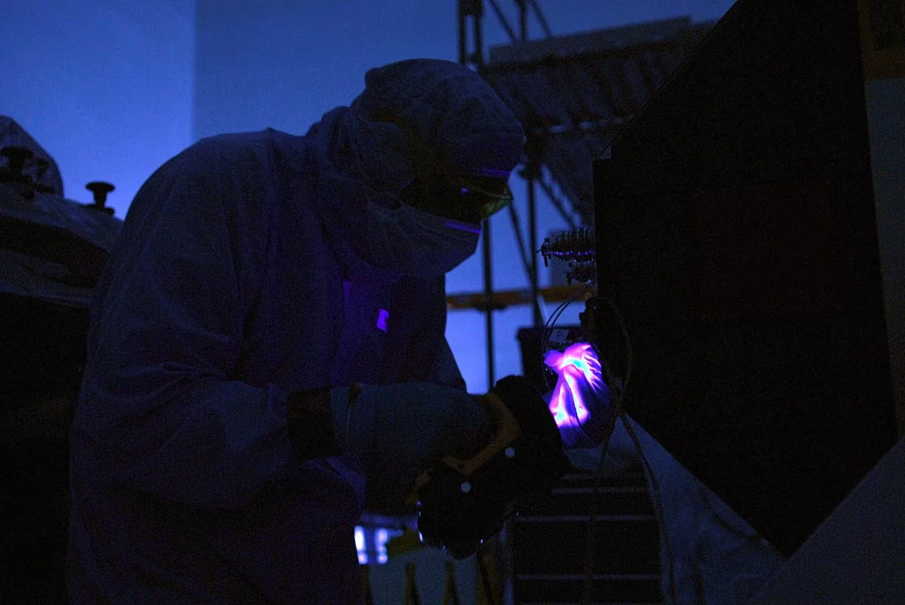 Clean-room technicians work beside equipment in dim blue illumination, with violet light near a gloved hand.