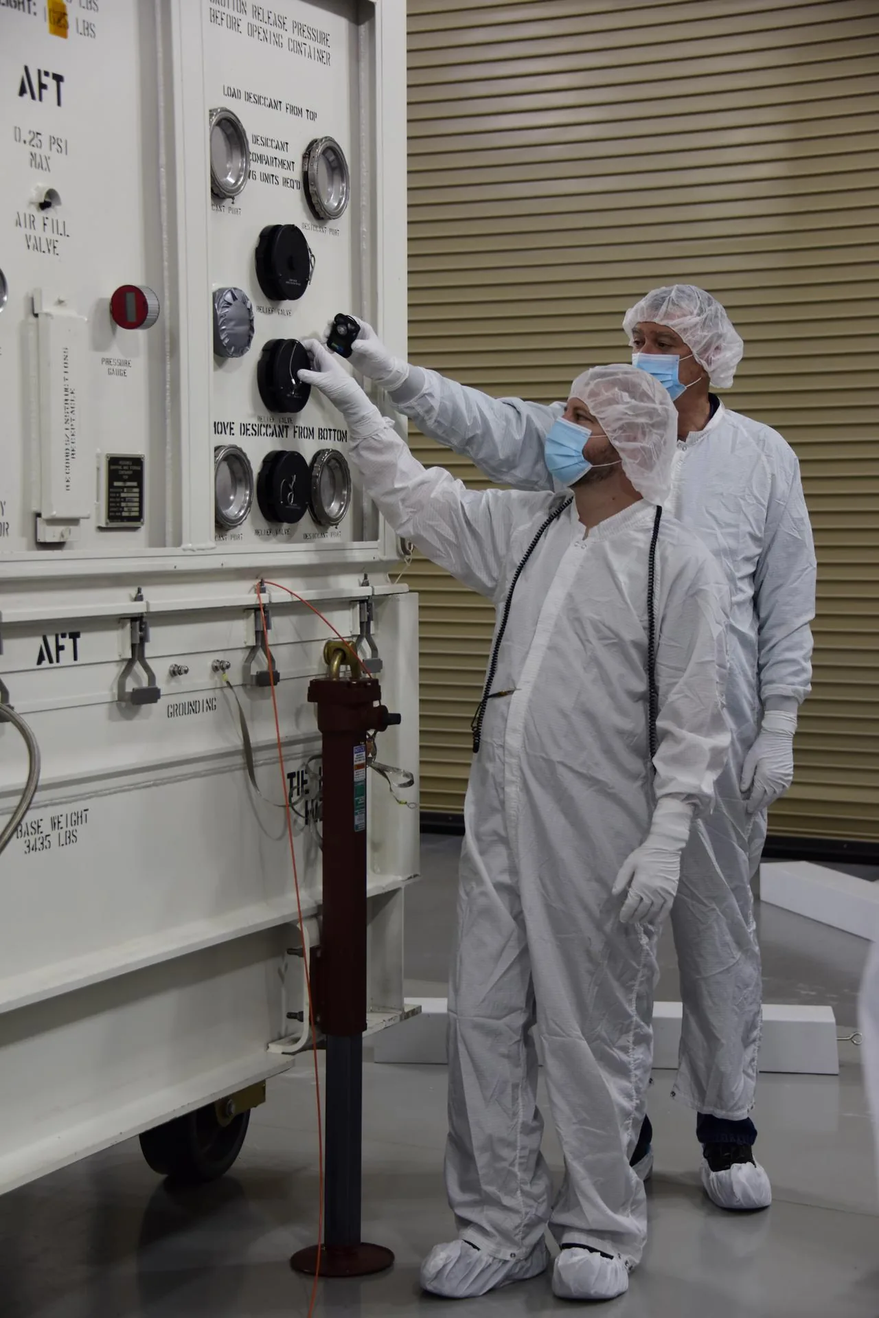 Two cleanroom technicians work at controls on a large shipping container