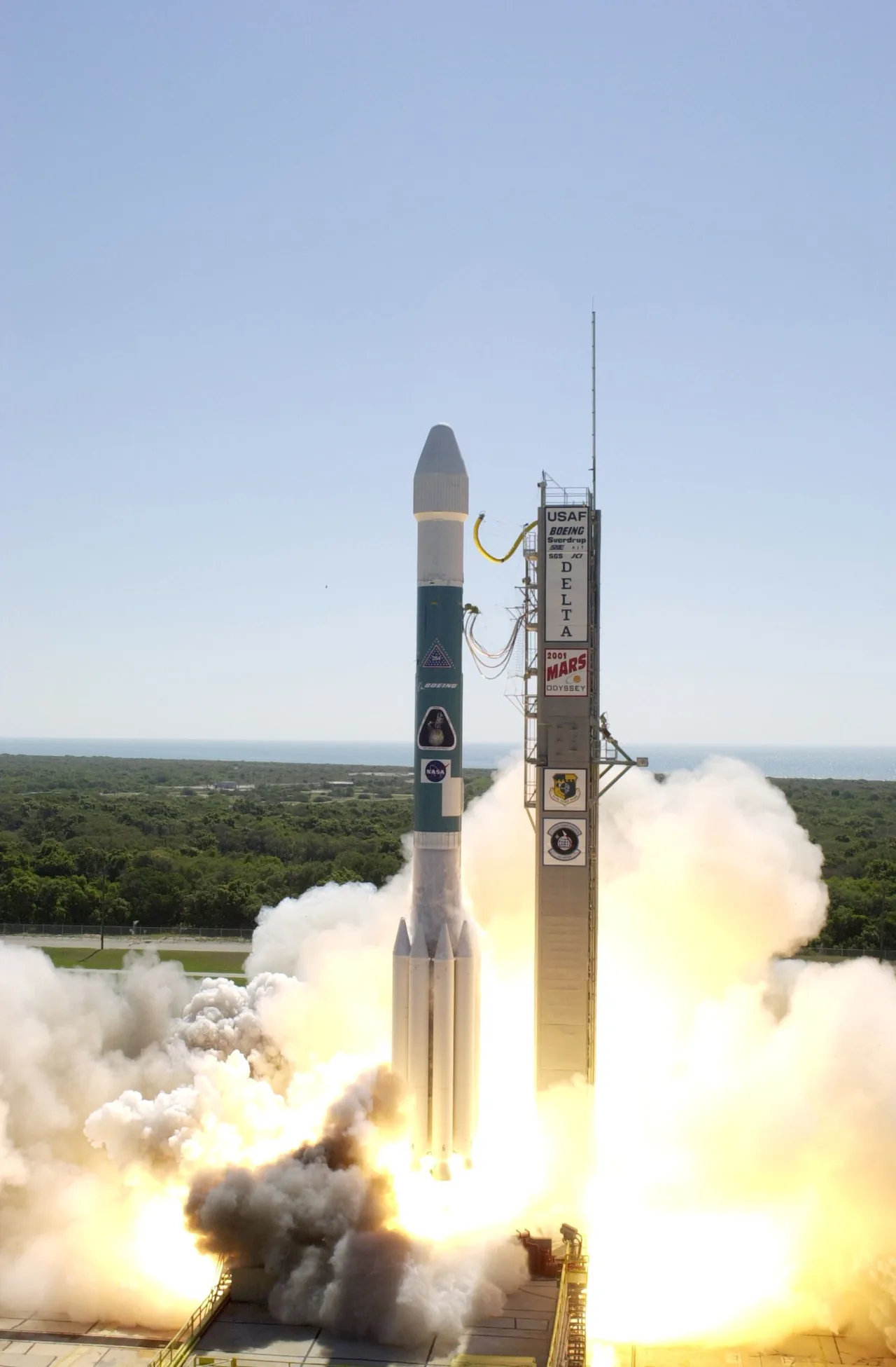 A blue-and-white Delta II lifts off beside a tower marked 2001 Mars Odyssey, above bright exhaust and smoke