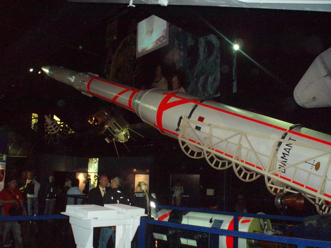 A white and red Diamant rocket is displayed horizontally above visitors in a dark museum gallery