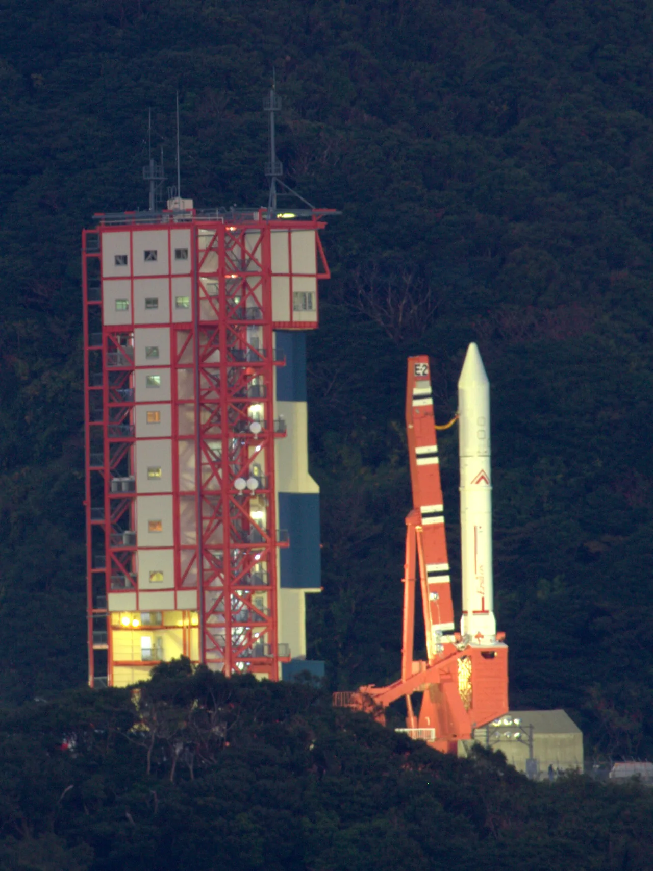 White Epsilon Flight 2 stands on a red launch mount beside an illuminated service tower, with wooded hills behind it.