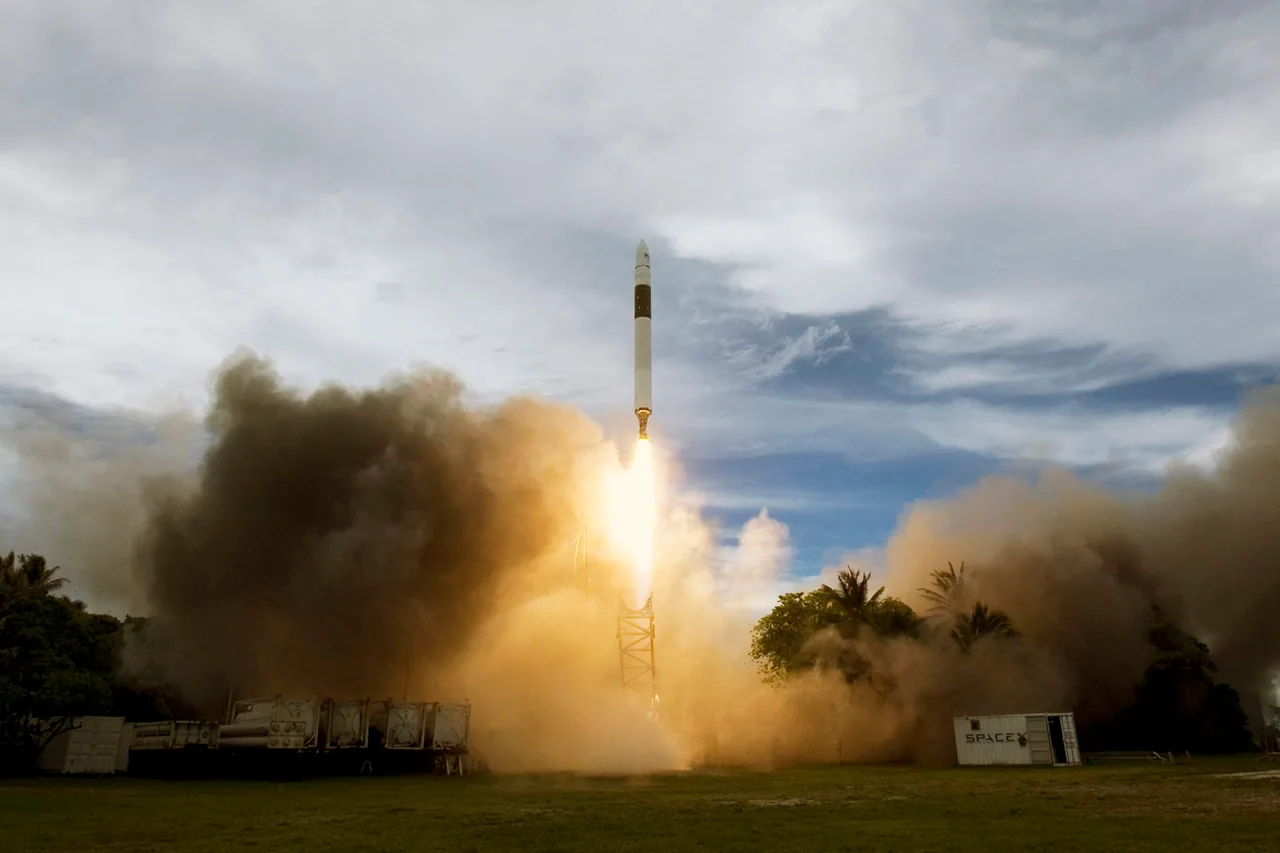 A slender Falcon 1 rises above palm trees and a low island launch site with a bright exhaust plume