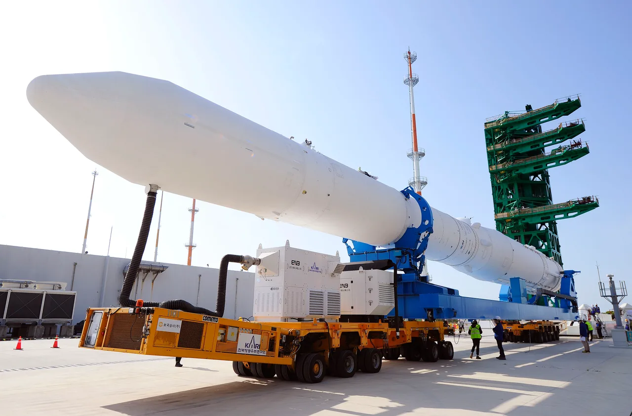 The white Nuri rocket lies horizontally on a blue and orange transporter beside a green launch tower