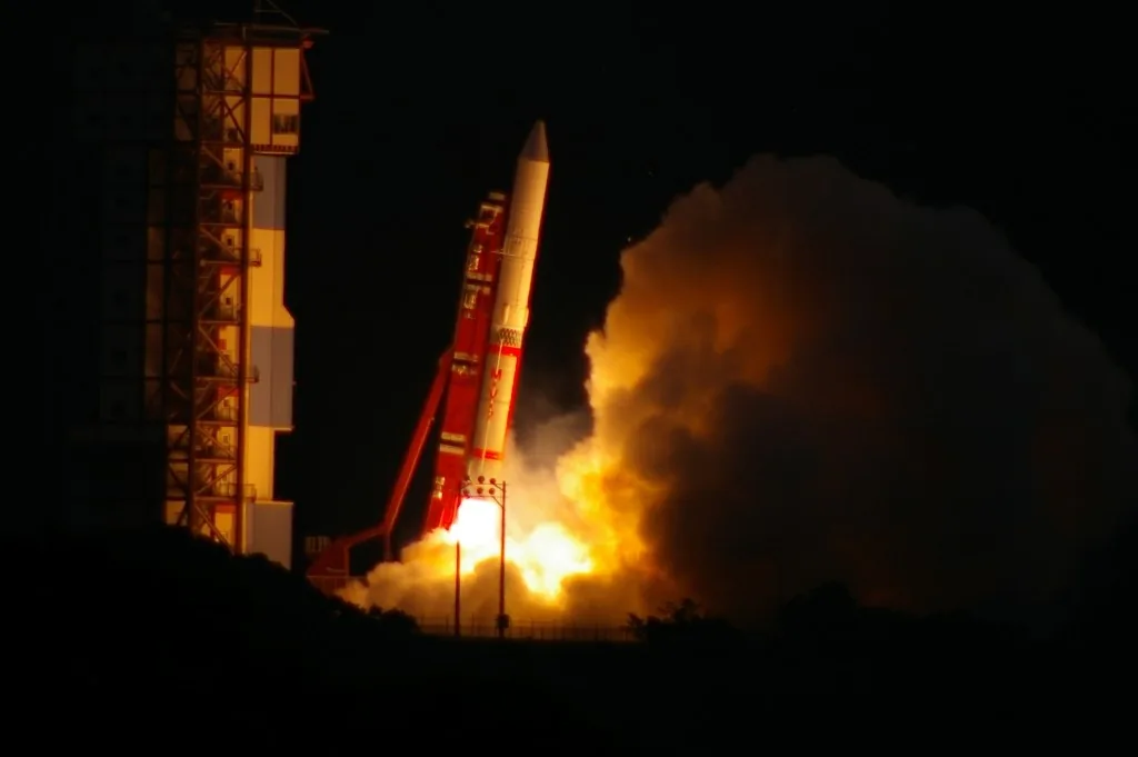 M-V No.7 rises beside its launch tower against a dark sky, with bright exhaust illuminating smoke below.