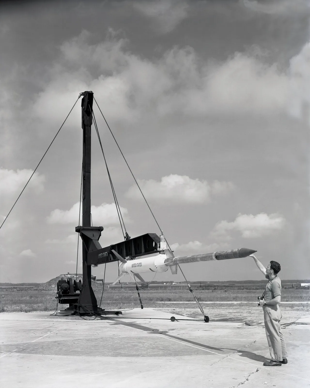 A sounding rocket sits horizontally on a launcher beside a technician holding its nose cover