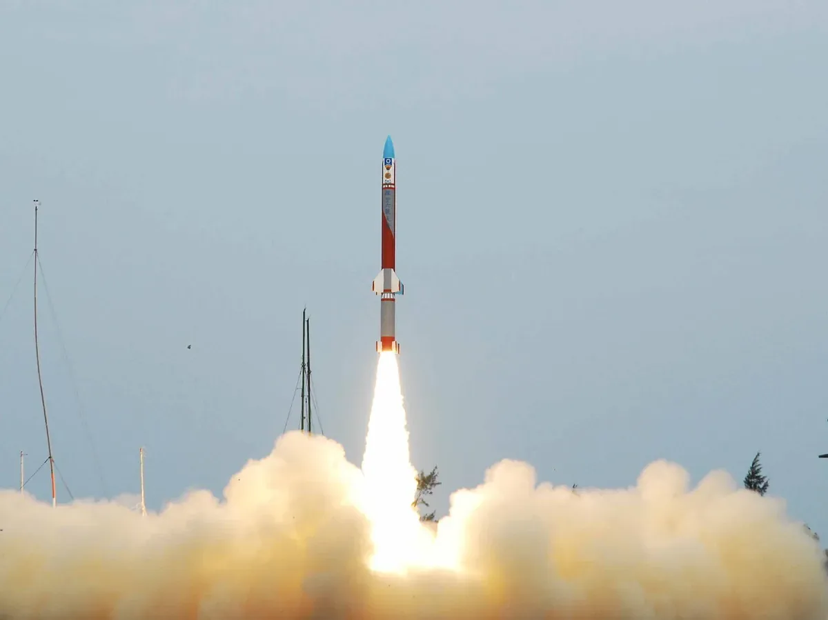 A red-and-white sounding rocket with a blue nose rises above a bright plume against a pale sky