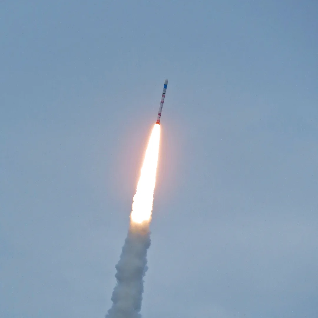 A slender SSLV rocket climbs above a bright flame and a broad smoke trail against a hazy blue sky