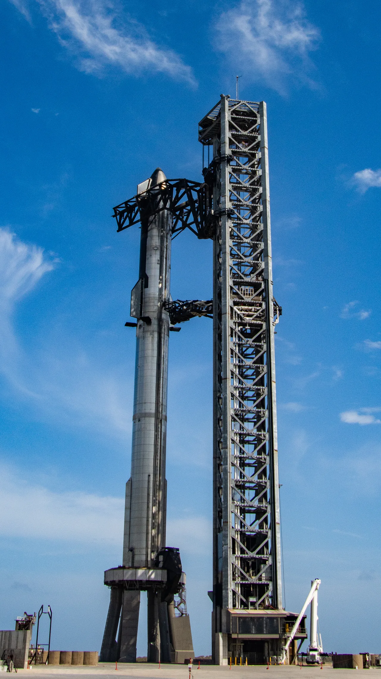 A complete stainless-steel Starship and Super Heavy stack stands vertically beside the launch tower under a vivid blue sky at SpaceX Starbase.