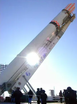 A white Zenit rocket is raised at an angle on its support structure, with people silhouetted below
